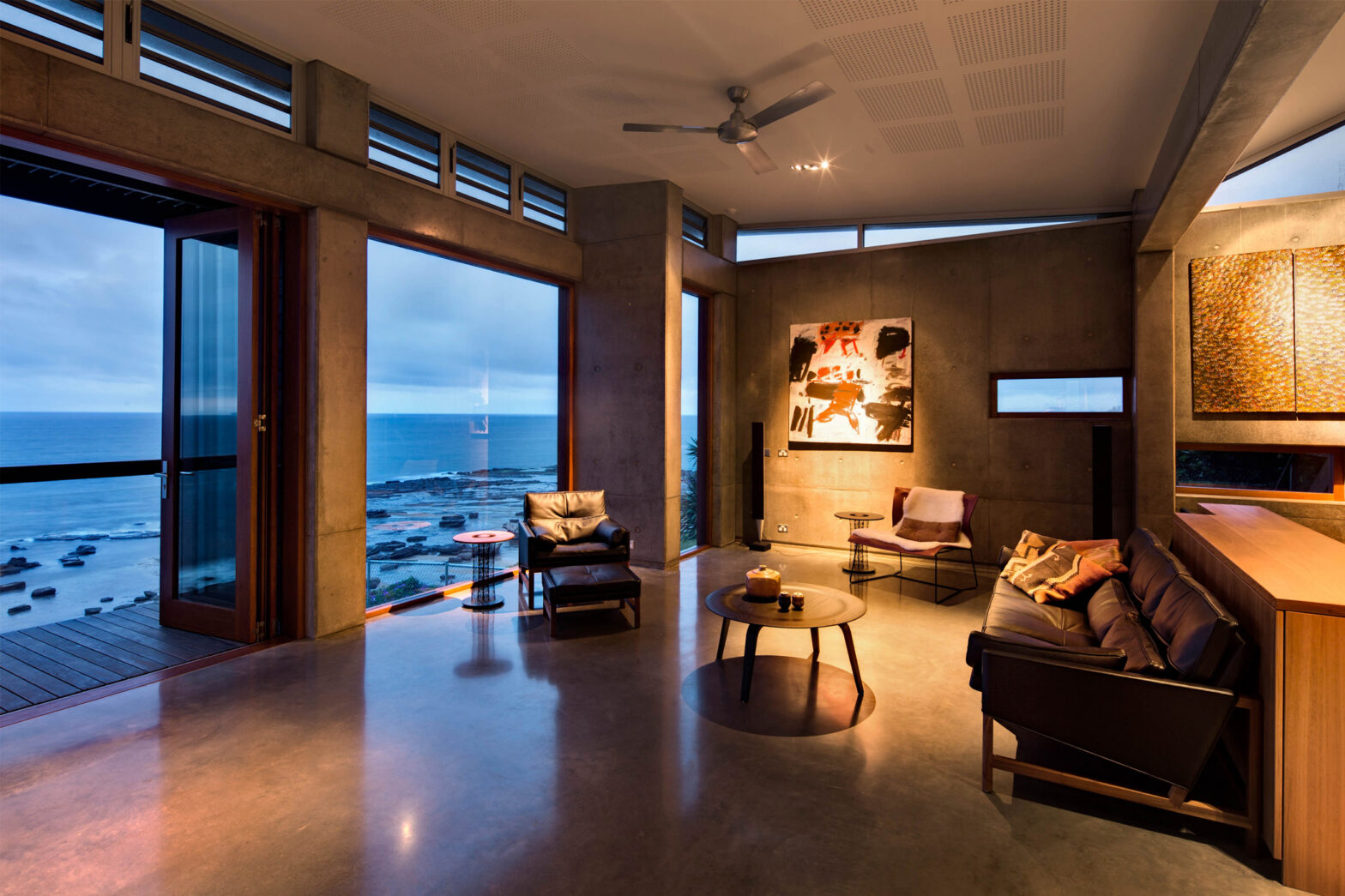 Modern living room with ocean view, open glass doors, abstract art, leather seating, and soft ambient lighting.