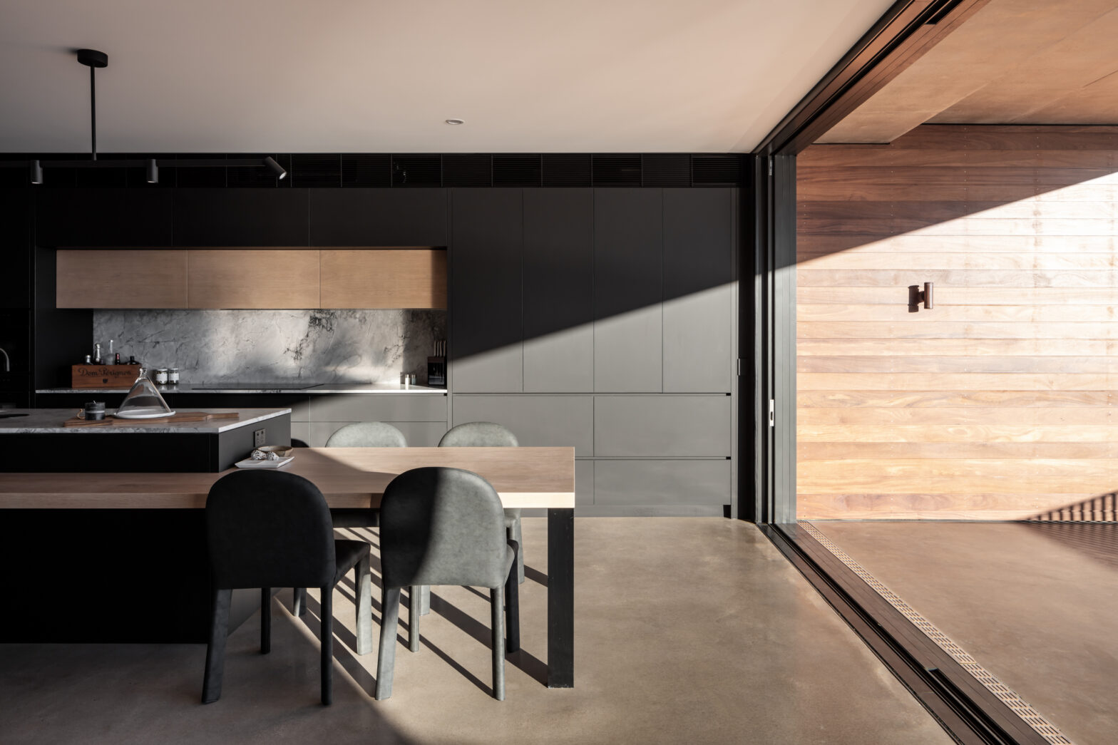 Modern kitchen with dark cabinets, a marble backsplash, wooden accents, and a large table with black chairs. Sunlight stre...