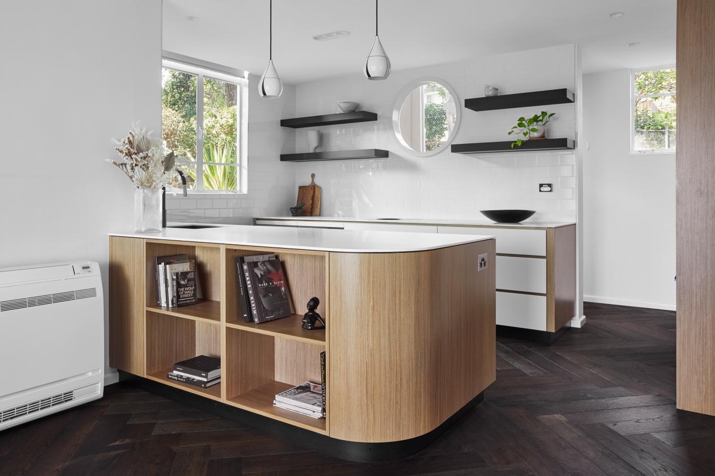 Modern kitchen with white cabinets, wooden island with books, black shelves, and a round window. Dark herringbone floor.