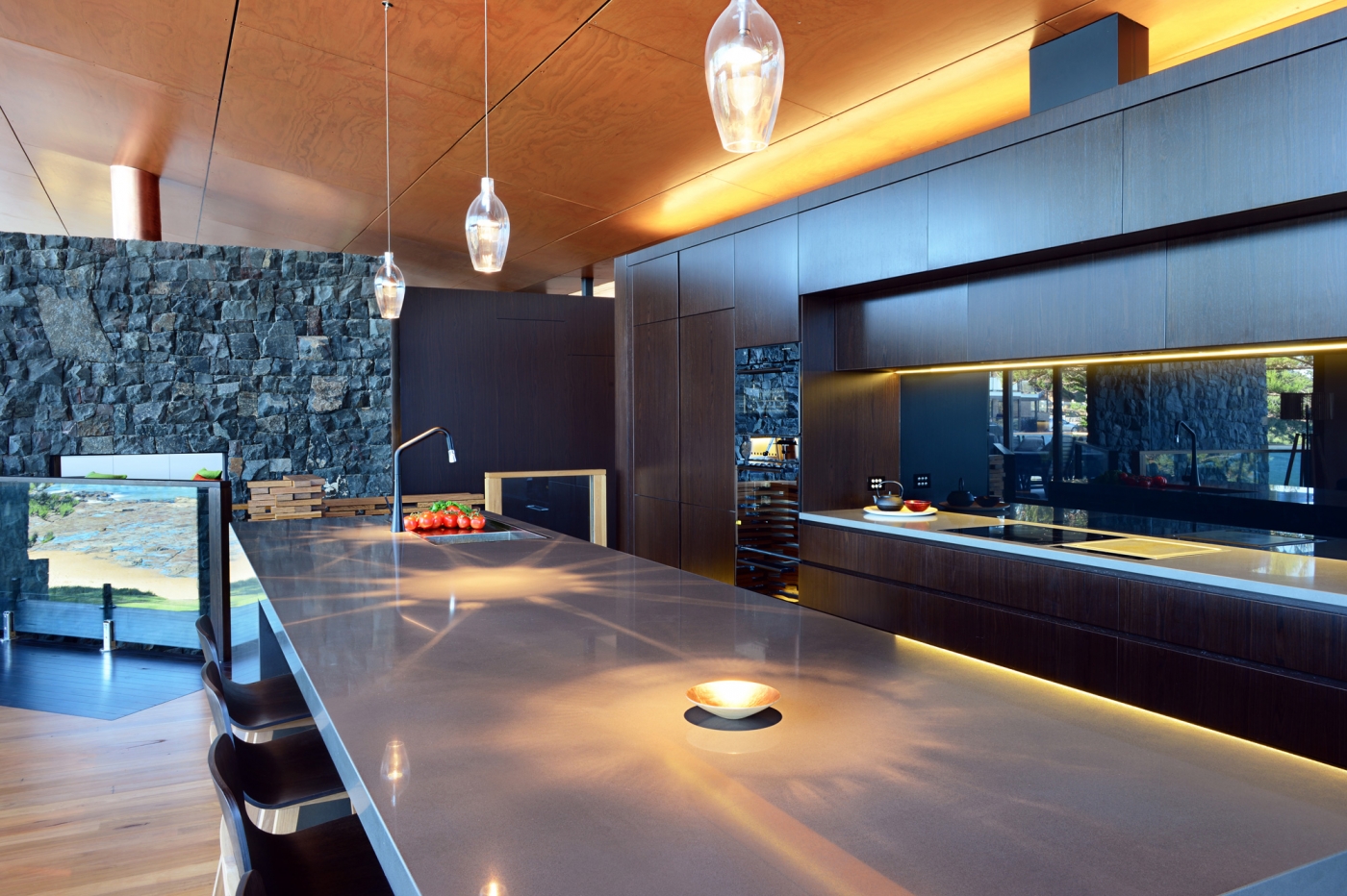 Modern kitchen with stone wall, sleek counters, pendant lights, and fruit bowl on an island. Warm wood and earthy tones.