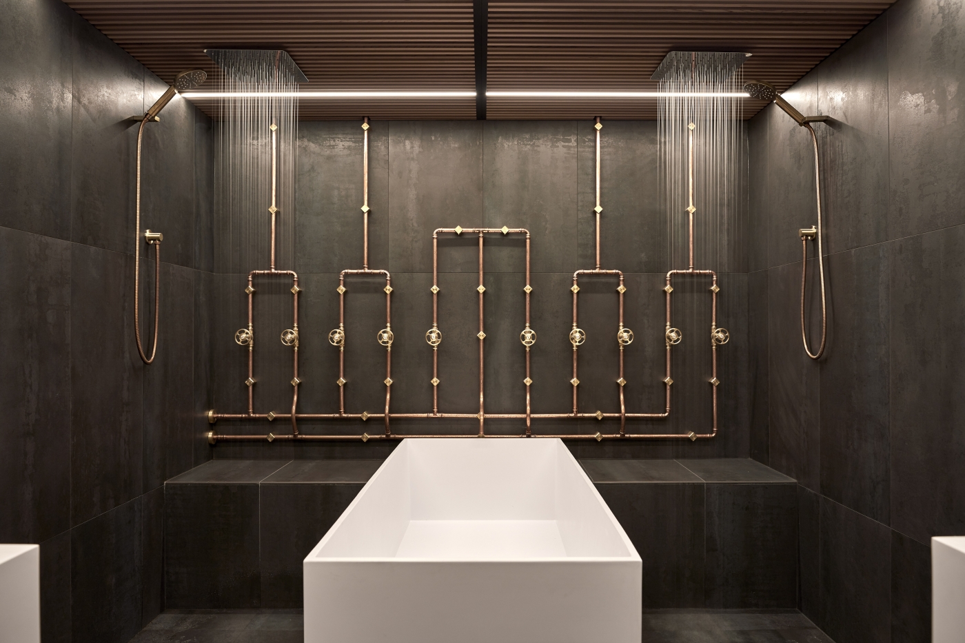 Modern bathroom with a freestanding white tub, copper pipe wall art, and dual rainfall showerheads on dark tiled walls.