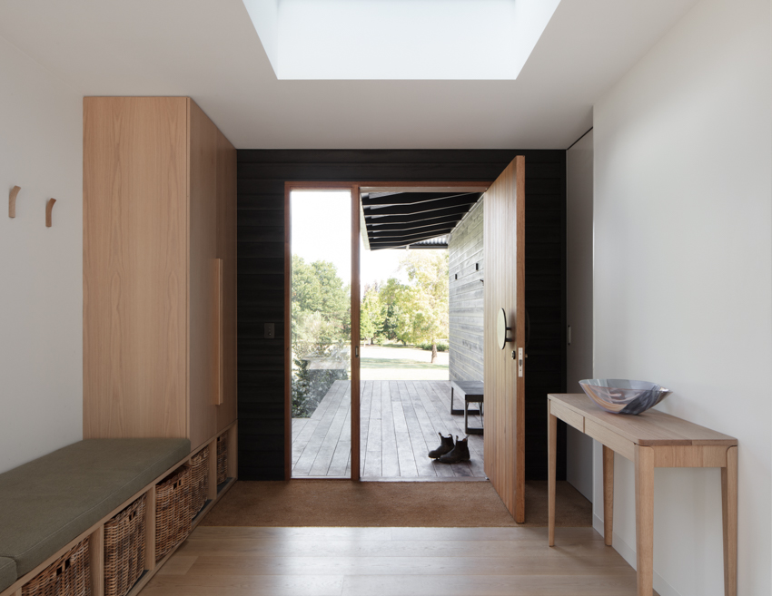 Modern entryway with wooden bench, skylight, and open door leading to a sunlit deck and green garden view.