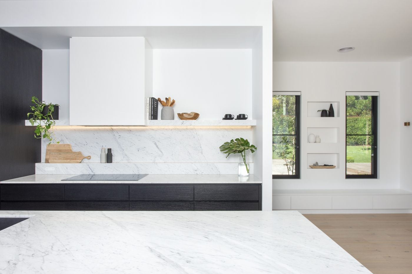Modern kitchen with white marble countertops, dark cabinets, floating shelf, and large windows overlooking green lawn.