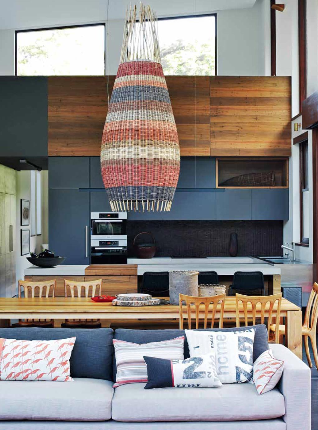 Modern living room and kitchen with striped chandelier, wooden cabinets, dining table, gray sofa, and colorful cushions.