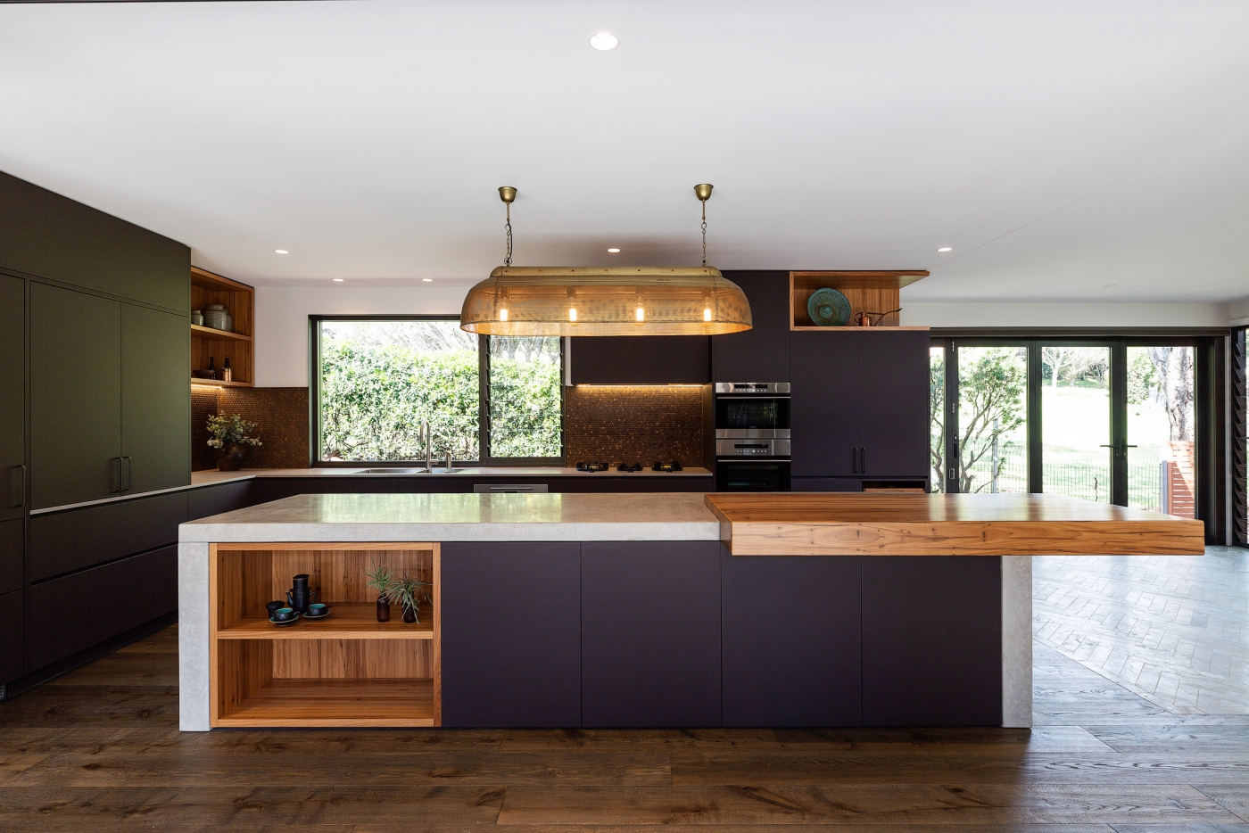 Modern kitchen with dark cabinets, large island, wooden accents, and brass pendant light, overlooking a garden through gla...