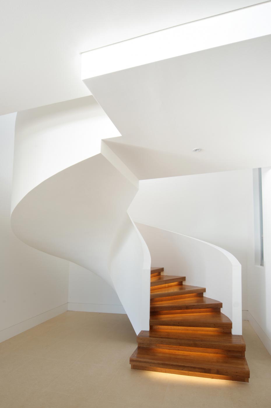 Spiral staircase with wooden steps and white curved railing, softly lit, in a modern, minimalistic interior.