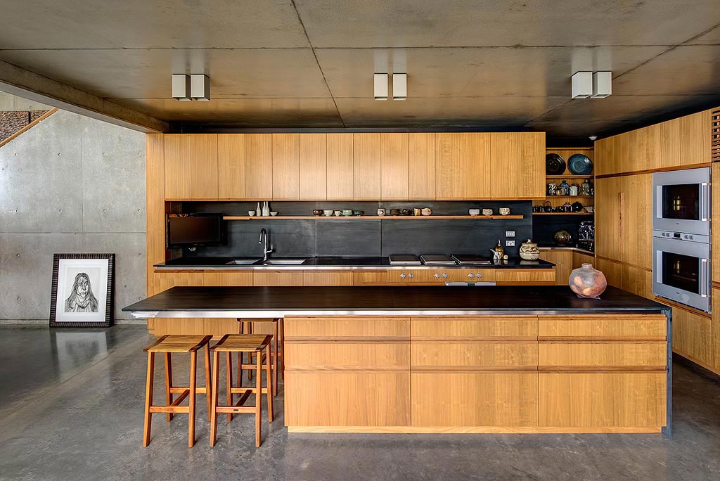 Modern kitchen with wooden cabinets, black countertops, and a central island with stools, framed art on concrete wall.