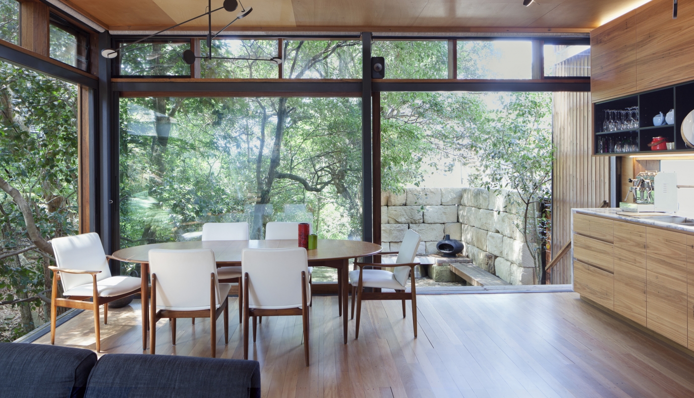 Modern dining room with glass walls, round table, white chairs, and view of lush greenery and large stone wall outside.