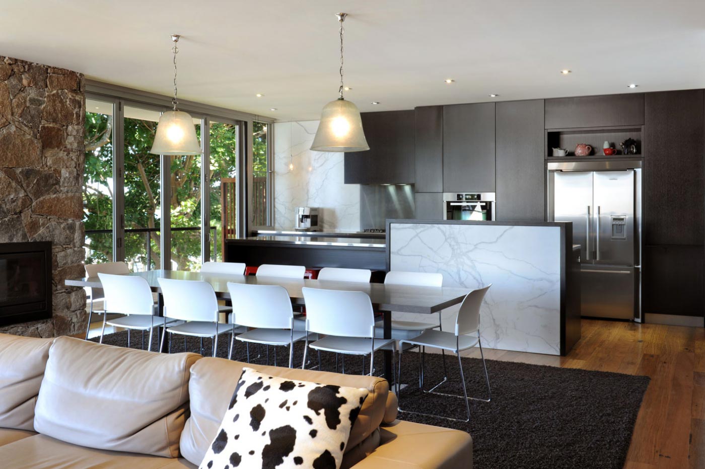 Modern kitchen and dining area featuring dark cabinets, marble island, white chairs, and pendant lights, with large windows.