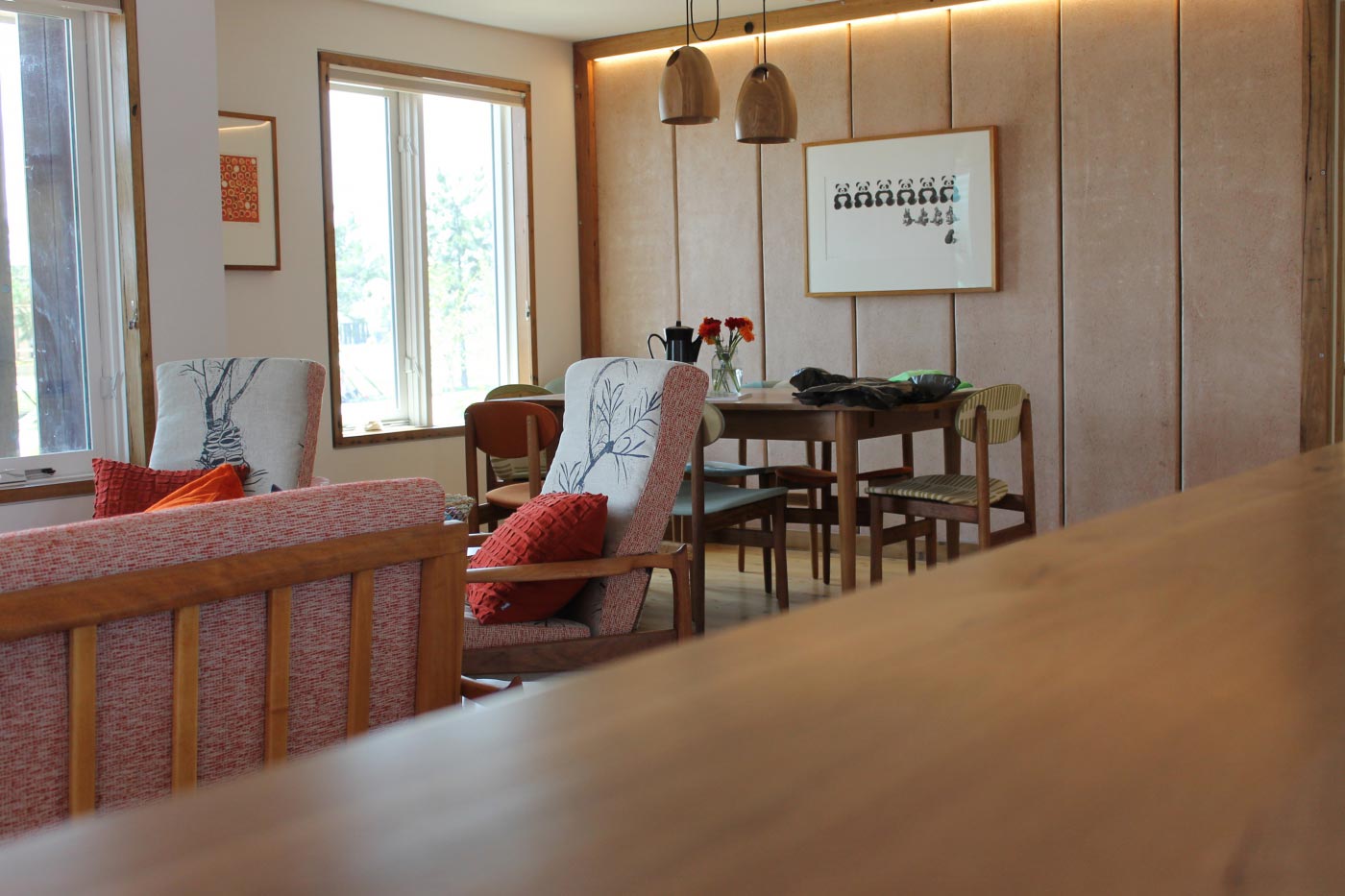 Cozy living room with red cushions, wooden furniture, and a dining area. Sunlight filters through large windows.