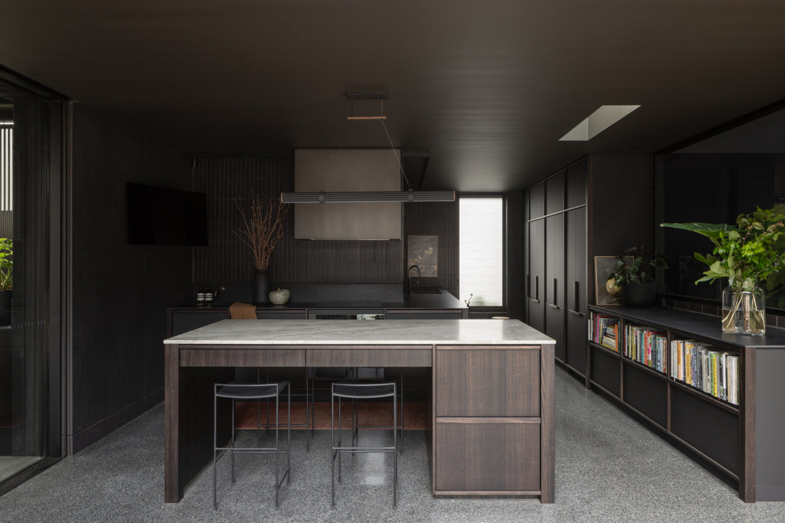 Modern kitchen with dark cabinetry, marble-top island, sleek bar stools, and plants on bookshelves.