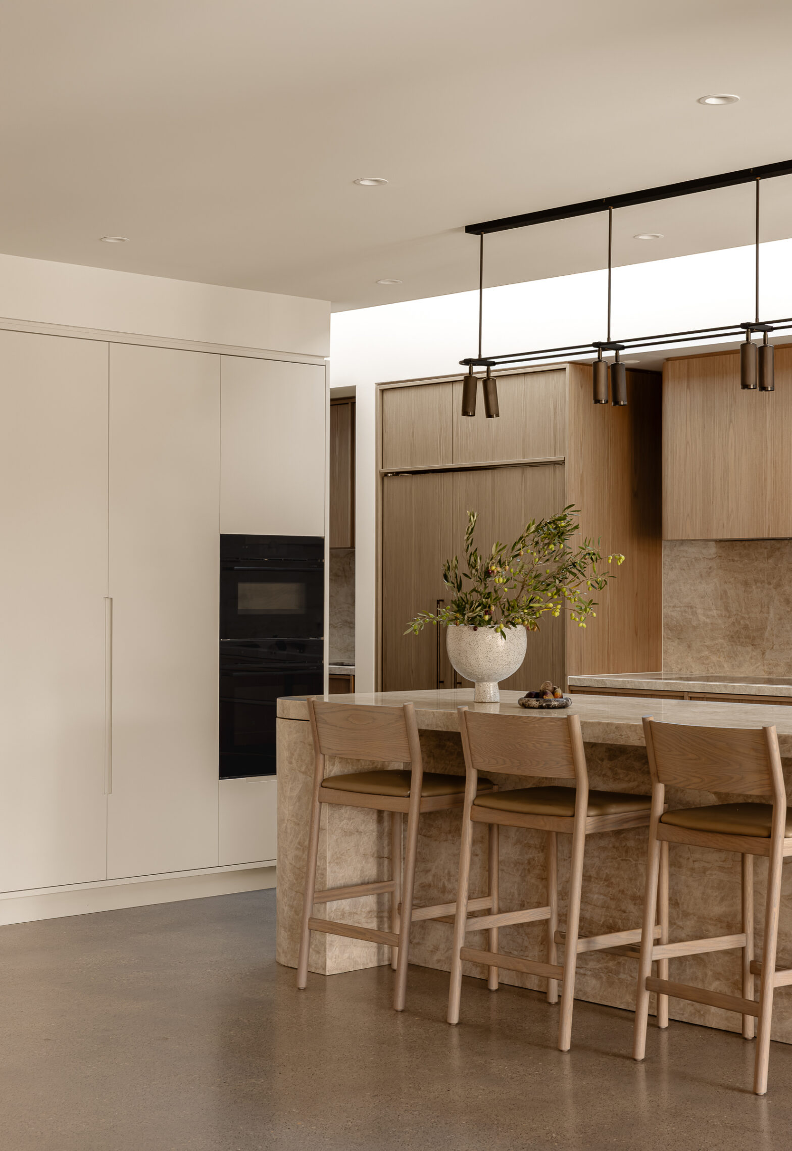Modern kitchen with marble island, four wooden chairs, tall vase with green branches, pendant lights, and wood cabinets.