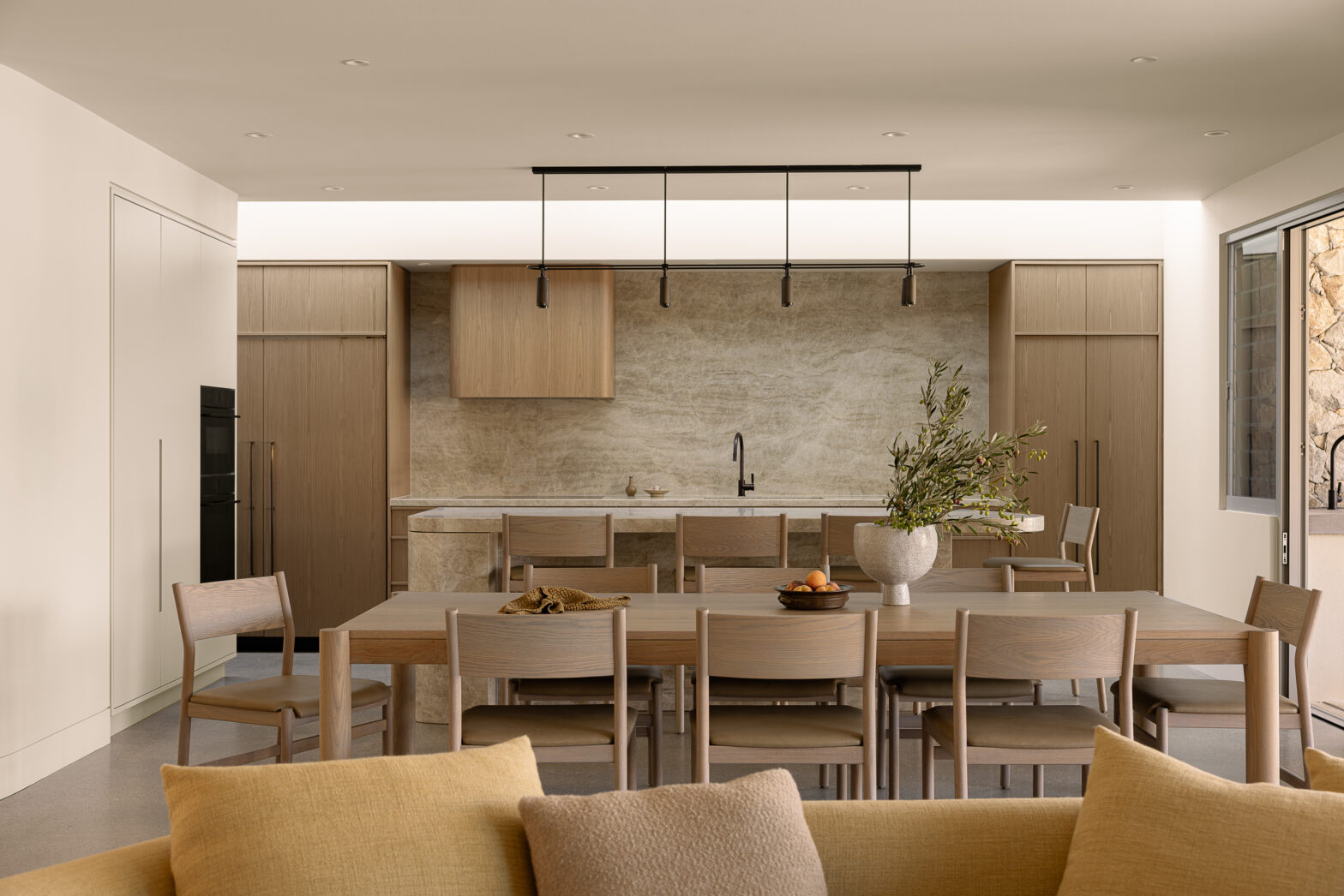 Modern kitchen with light wood cabinets, marble backsplash, long dining table, and minimalist decor.