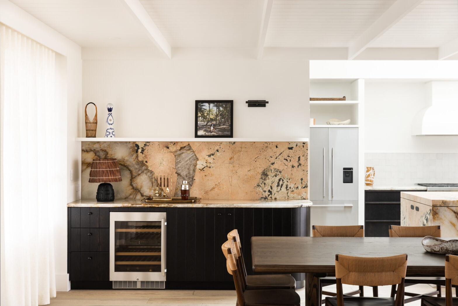 Modern kitchen with dark cabinets, marble backsplash, and wooden dining table. Wine fridge and decorative lamp on countertop.